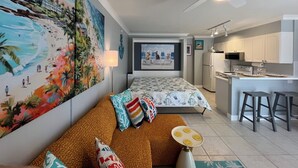 Een modern appartement met een strandmuur, een bank met kleurrijke kussens, een eethoek met krukken, en een keuken met witte kasten.