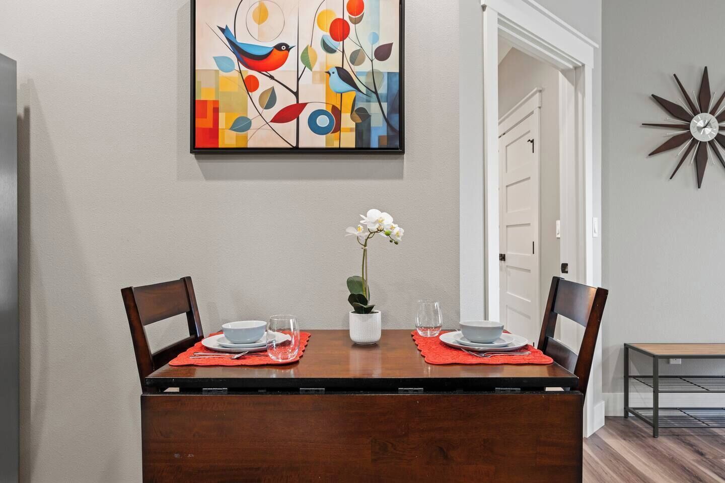 Een eetkamer met een houten tafel, twee stoelen, een vaas met bloemen en een kleurrijk abstract schilderij aan de muur.