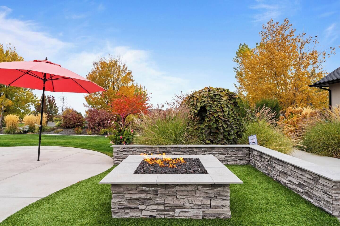 Un espace extérieur aménagé avec un foyer en pierre, un parasol rouge et divers arbustes et arbres.