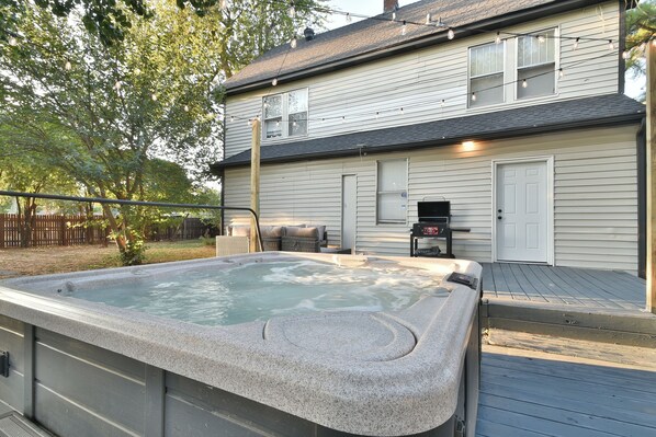 Ein Whirlpool in einem Garten mit Holzterrasse, Grill und einem Haus mit weißer Fassade.