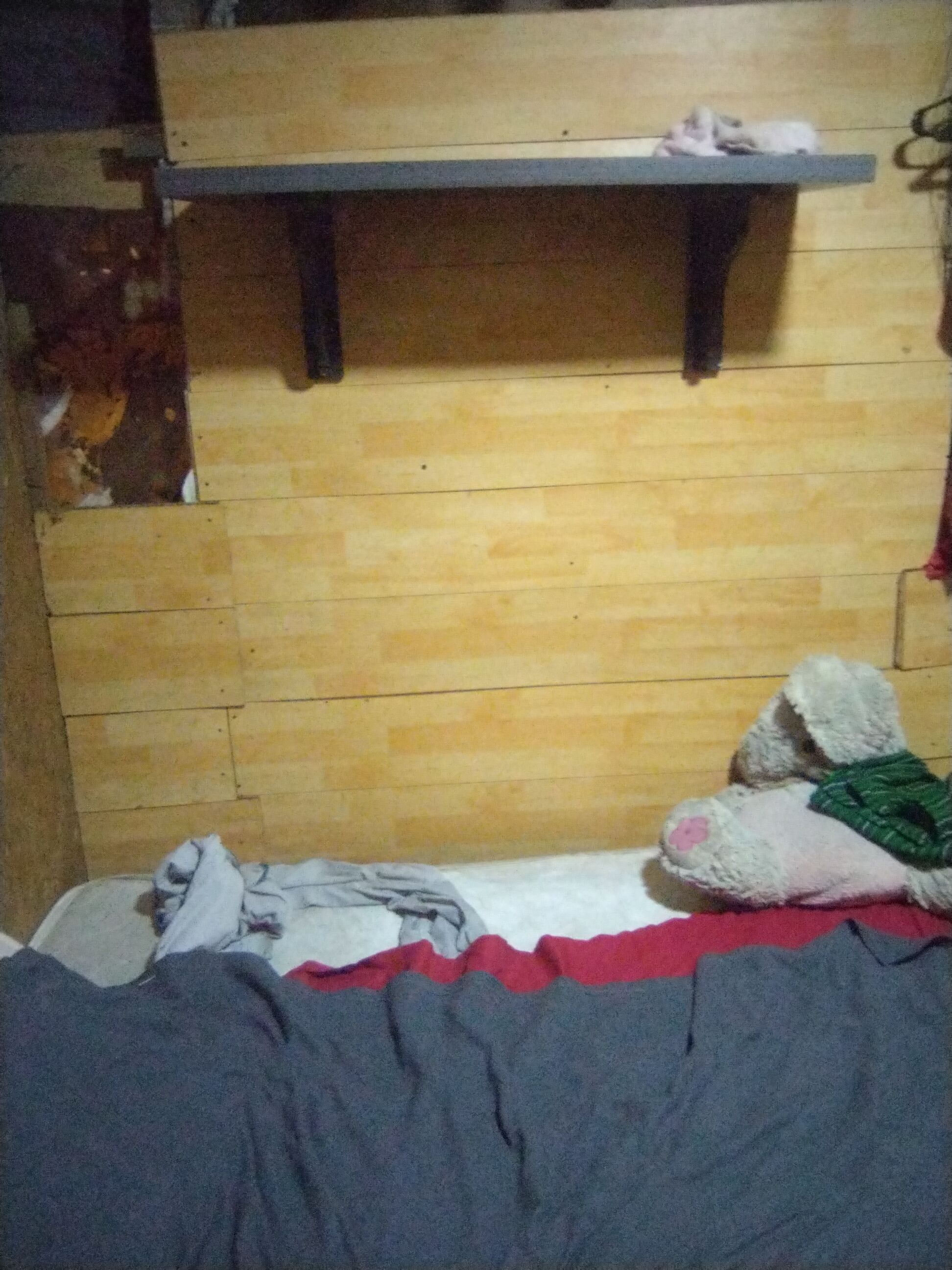 Una habitación pequeña, con poca luz, que tiene una cama individual, un estante de madera encima y una pared con un diseño de azulejos.