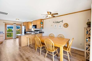 Een moderne keuken met houten kastjes, een eethoek met een houten tafel en stoelen, een plafondventilator en uitzicht op een blauw huis door een schuifdeur.