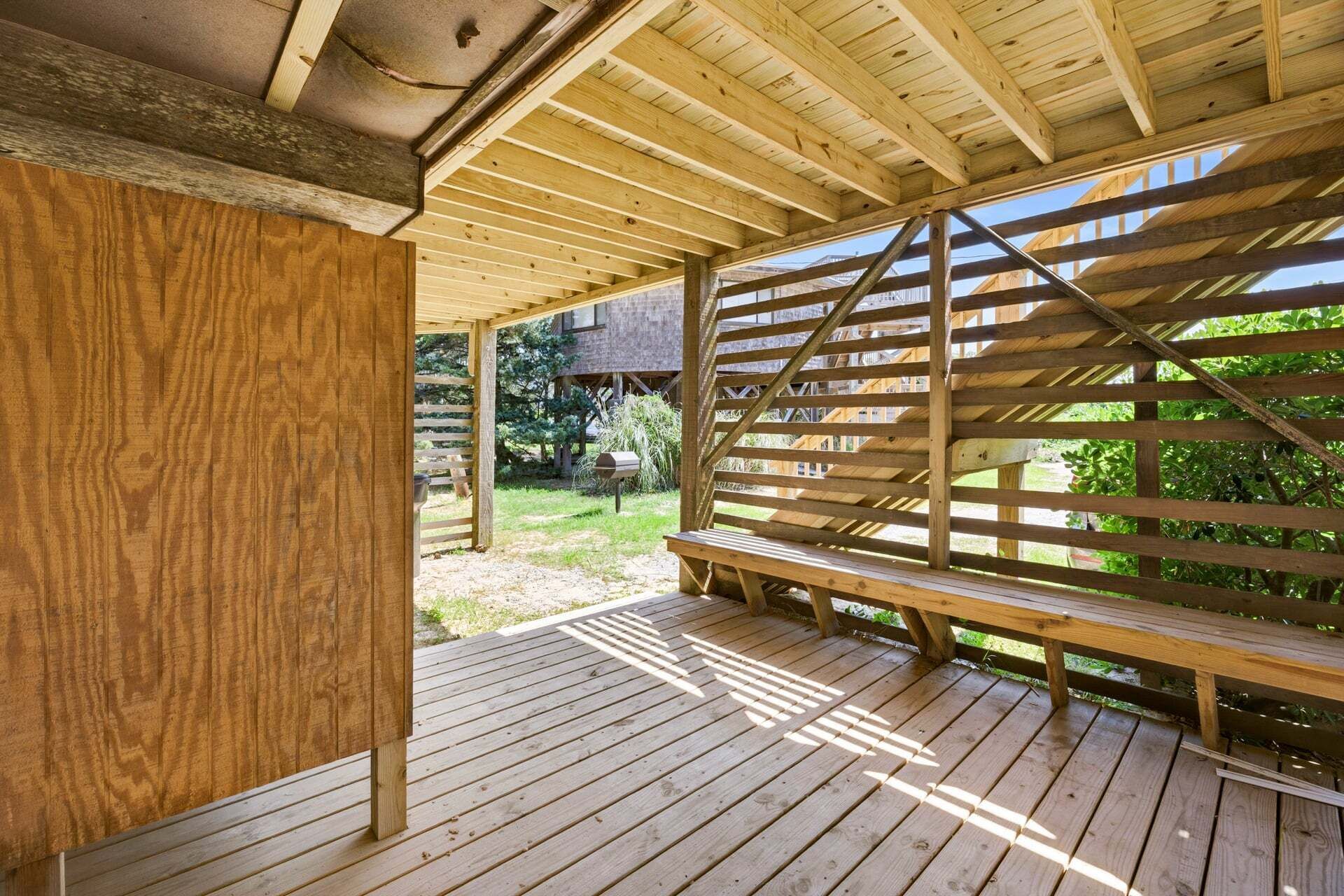 Una terraza de madera al aire libre con un banco, una pérgola y vistas a un jardín y un edificio.