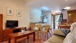 Un salon avec un téléviseur, une table basse, des fauteuils en osier, un canapé et, au fond, une cuisine.