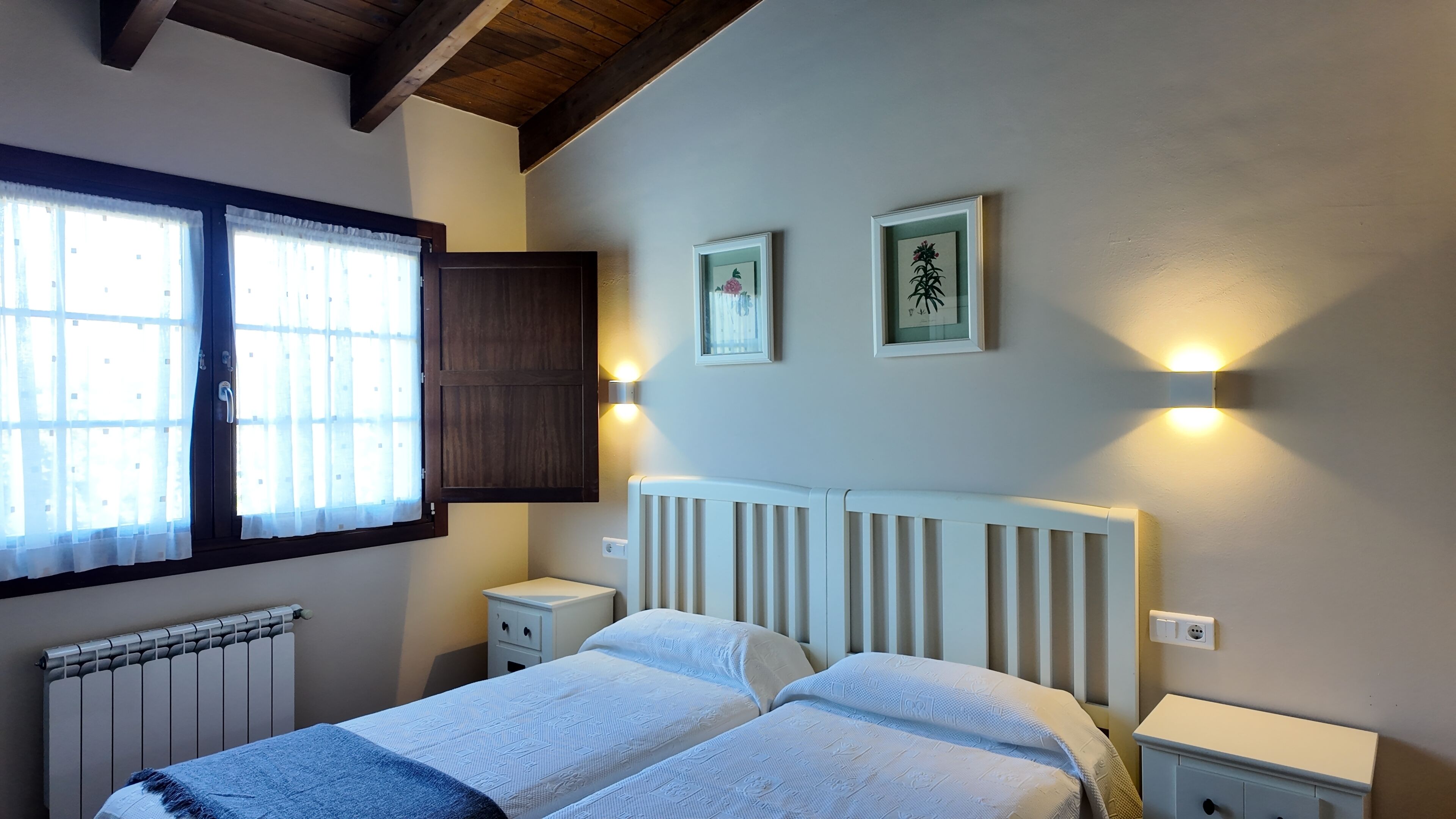 Ein Schlafzimmer mit zwei Betten, einem Fenster mit Vorhängen, einem Heizkörper und Wandleuchten.