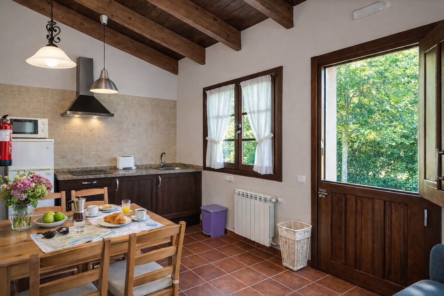 Une cuisine chaleureuse avec des poutres en bois, une table à manger dressée pour le petit-déjeuner et une fenêtre ornée de rideaux.