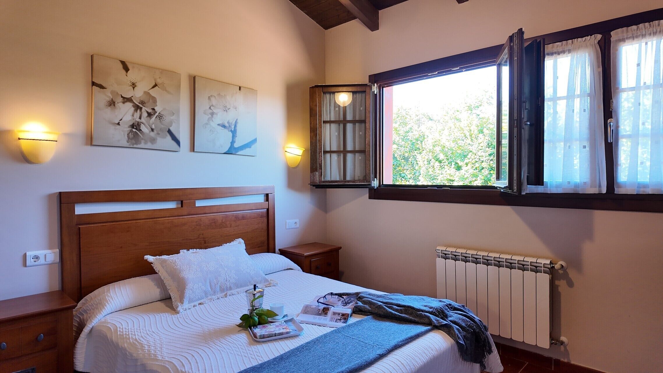 A bedroom with a wooden bed, a nightstand, a bedside table, a window with curtains, and wall-mounted artwork.