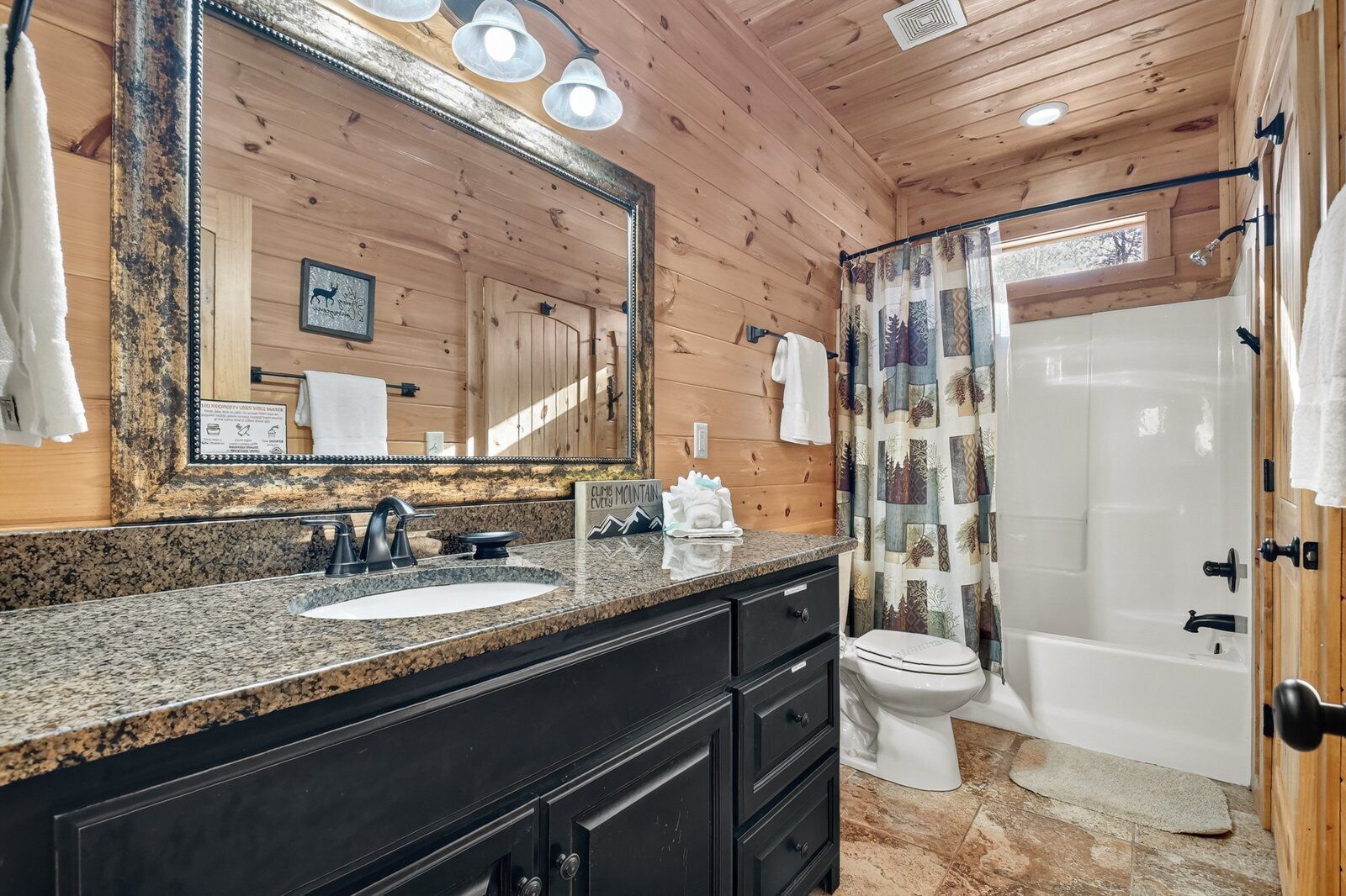Une salle de bain rustique, avec des murs en bois, un lavabo sur un plan de travail en granit, un grand miroir, des toilettes et une douche avec un rideau.