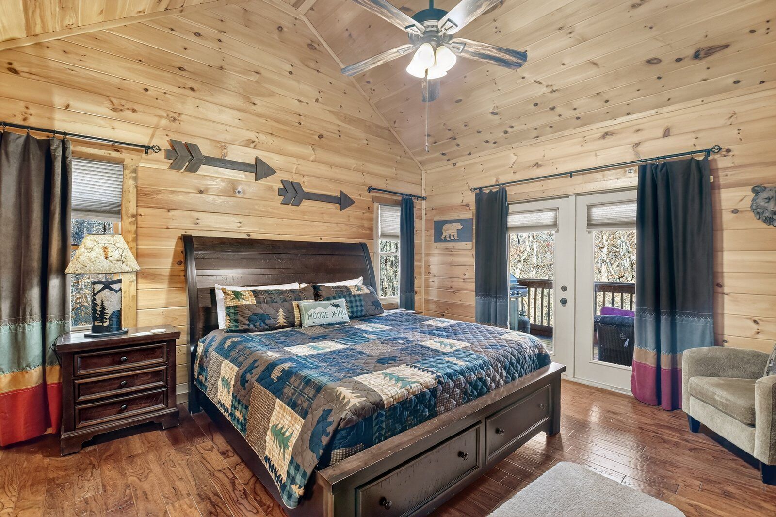 A cozy bedroom with a wooden ceiling, a bed with a quilt, a nightstand, a lamp, and a view of the outdoors.