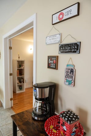A kitchen area with a coffee maker on a wooden table, a Mickey Mouse-themed basket, and various kitchen signs on the wall. - Bright and Cozy Cottage in the Heart of Dartmouth (Dartmouth)