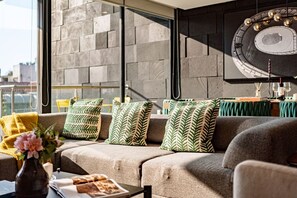 Una sala de estar moderna con un sofá, cojines estampados, una mesa de centro con un jarrón y una revista, y un ventanal con vistas a la ciudad.