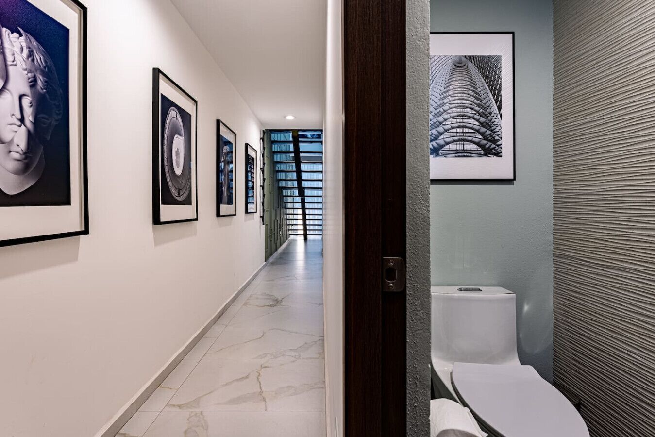 A hallway with framed artwork, a marble floor, and a view into a bathroom with a modern toilet and textured wall.