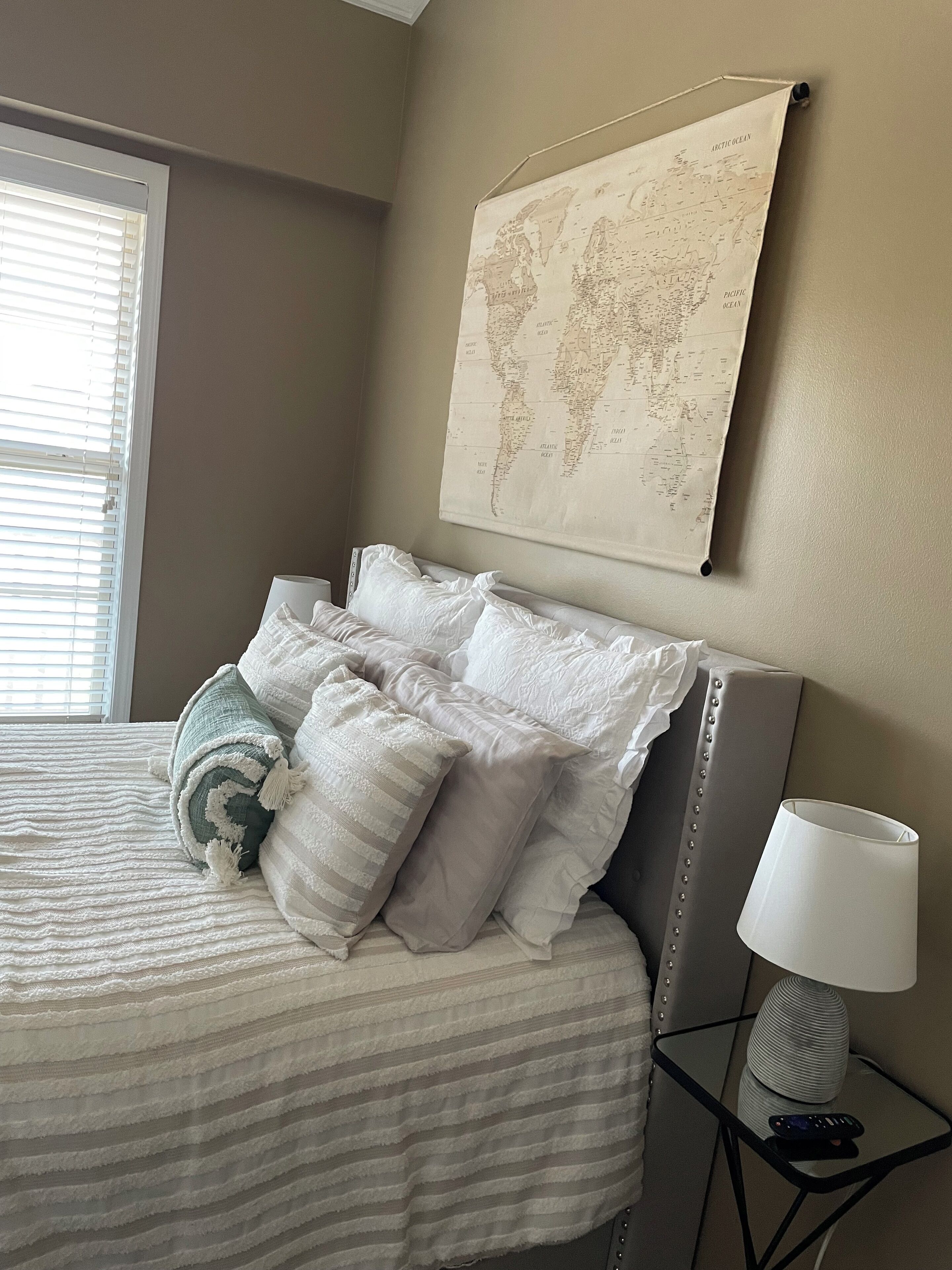 Una cama bien hecha con ropa de cama a rayas y varias almohadas, un mapa del mundo en la pared y una lámpara en una mesita de noche.