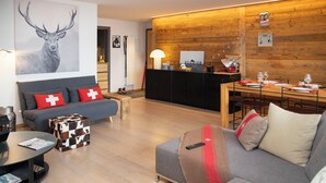 Ein modernes Wohnzimmer mit grauem Sofa, einem Holzesstisch für vier Personen und einer Küchenzeile mit Holzvertäfelung und einem schwarzen Schrank.