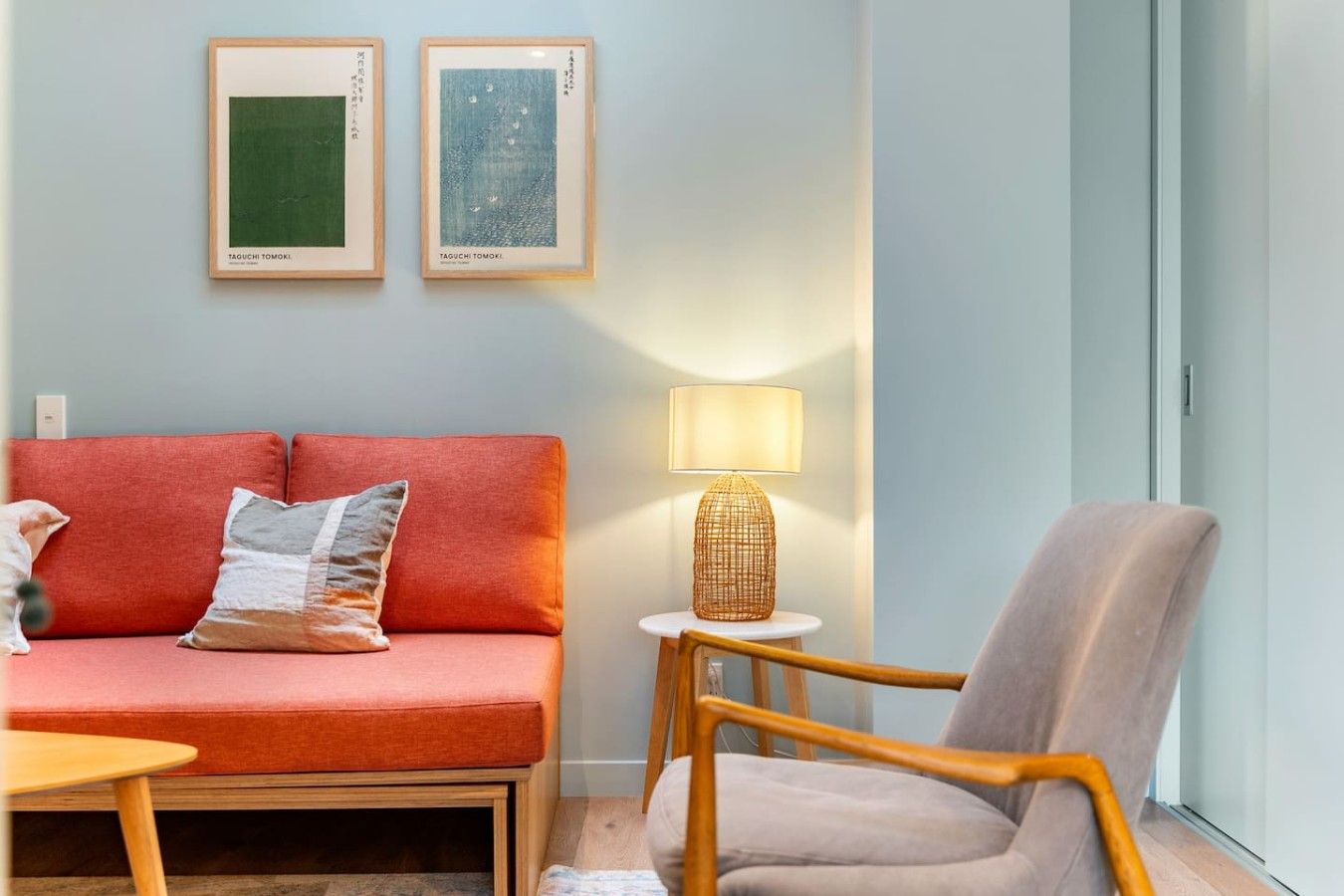 Een woonkamer met een rode bank, een grijze fauteuil, een kleine tafel en een lamp. Twee kunstwerken in lijsten hangen aan de muur.
