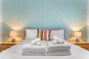 Una cama bien hecha con dos almohadas y una almohada a rayas, dos lámparas de noche y un par de toallas dobladas sobre una mesita de madera.