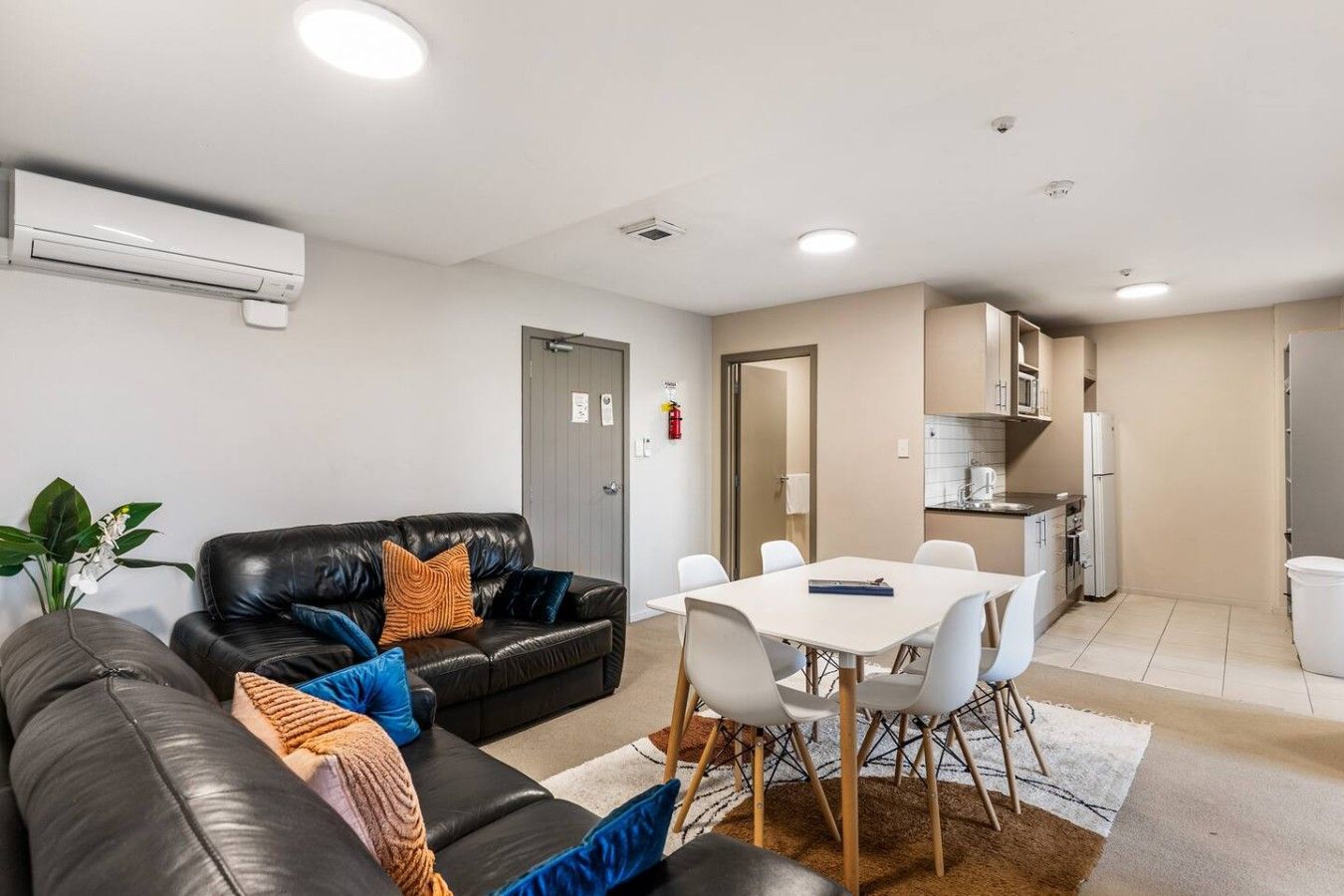 A modern living room with a black leather sofa, white dining table, and a kitchen area with white appliances.