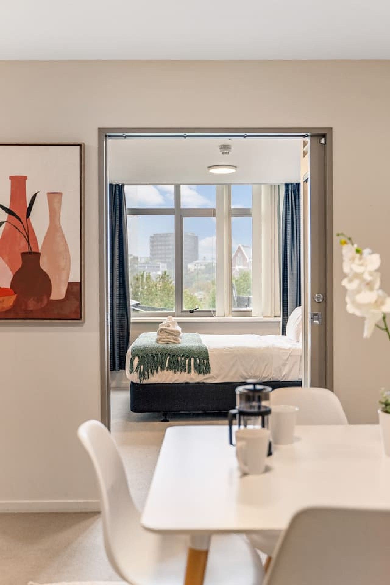 A hotel room with a bed, a table, and a chair. There is a window with curtains, and a painting on the wall.