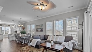 A spacious living room with a large sectional sofa, a dining area with a white table and chairs, and a kitchen with a view of the beach.