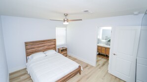 Quarto com cama de madeira, mesa de cabeceira, ventilador de teto, banheiro com pia e espelho, e porta branca.