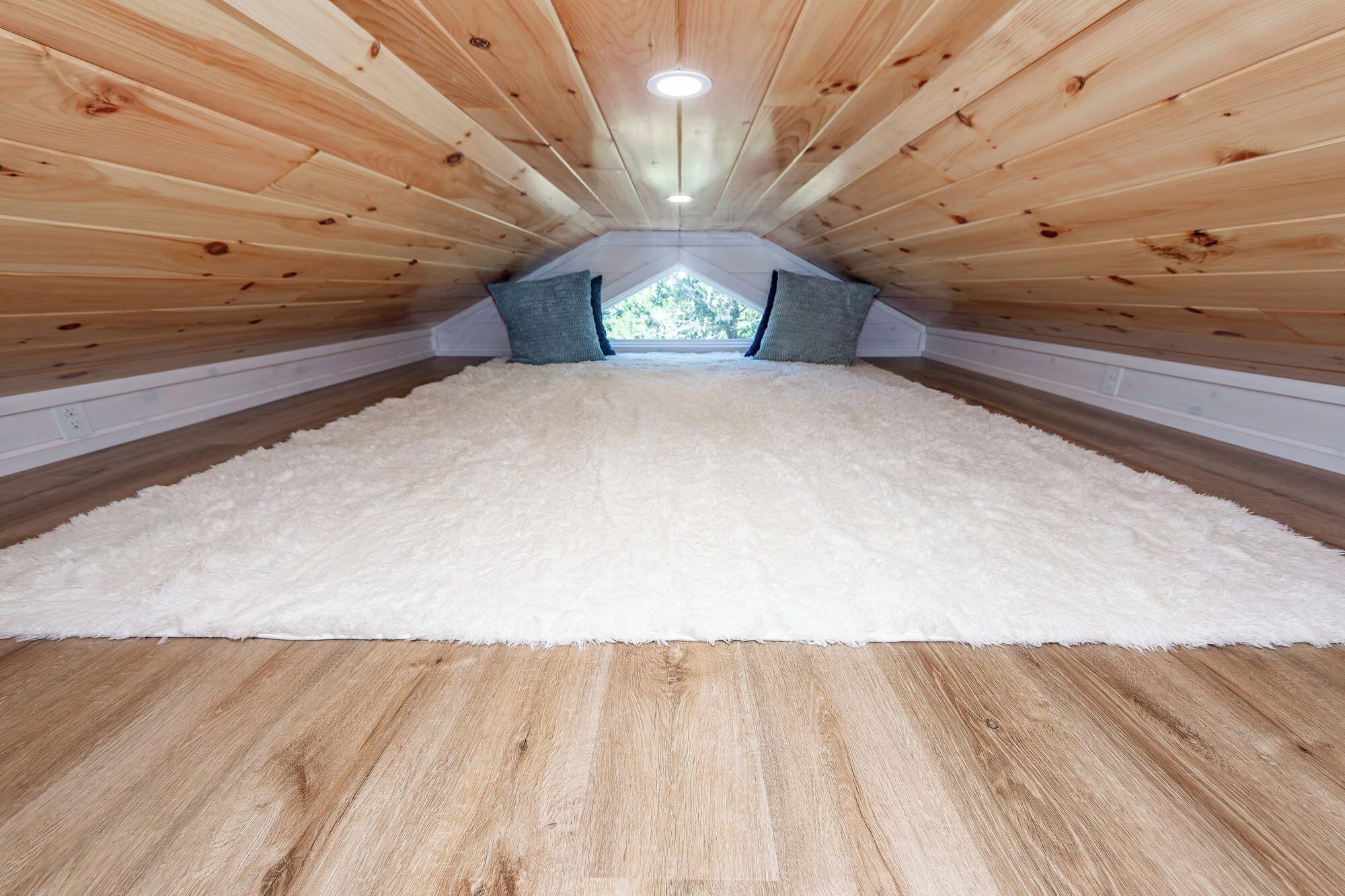 A room with a sloped wooden ceiling, a white fluffy rug, and a view through a window.