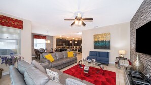 Uma sala de estar moderna com um sofá cinzento, um tapete vermelho e uma poltrona azul. Há um ventilador de teto, um quadro na parede e uma TV à direita.