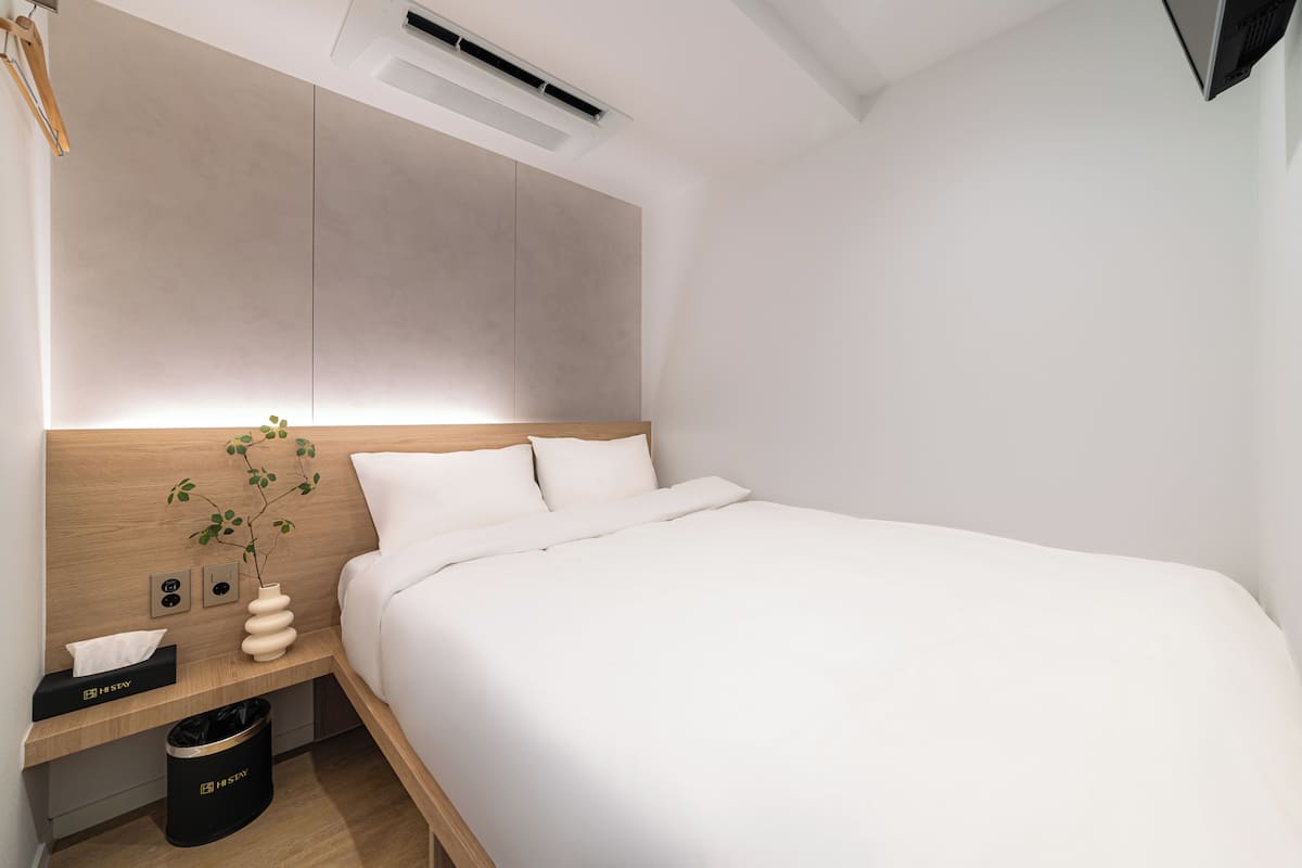 A modern hotel room with a wooden headboard, a bedside table with a vase of green leaves, a black trash bin, and a white bedspread.