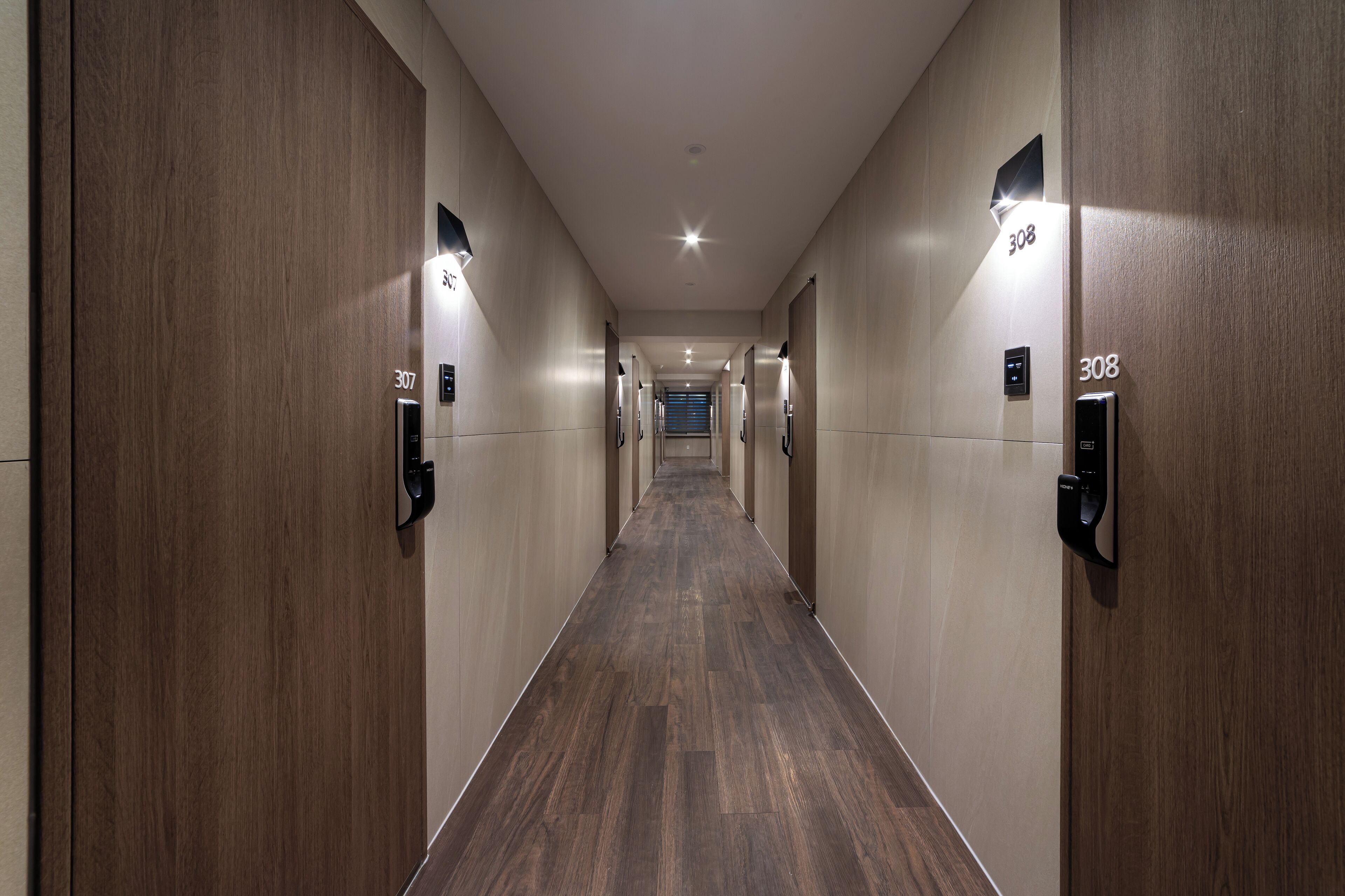 A long, narrow hallway with wooden paneling and numbered doors, leading to a distant exit.