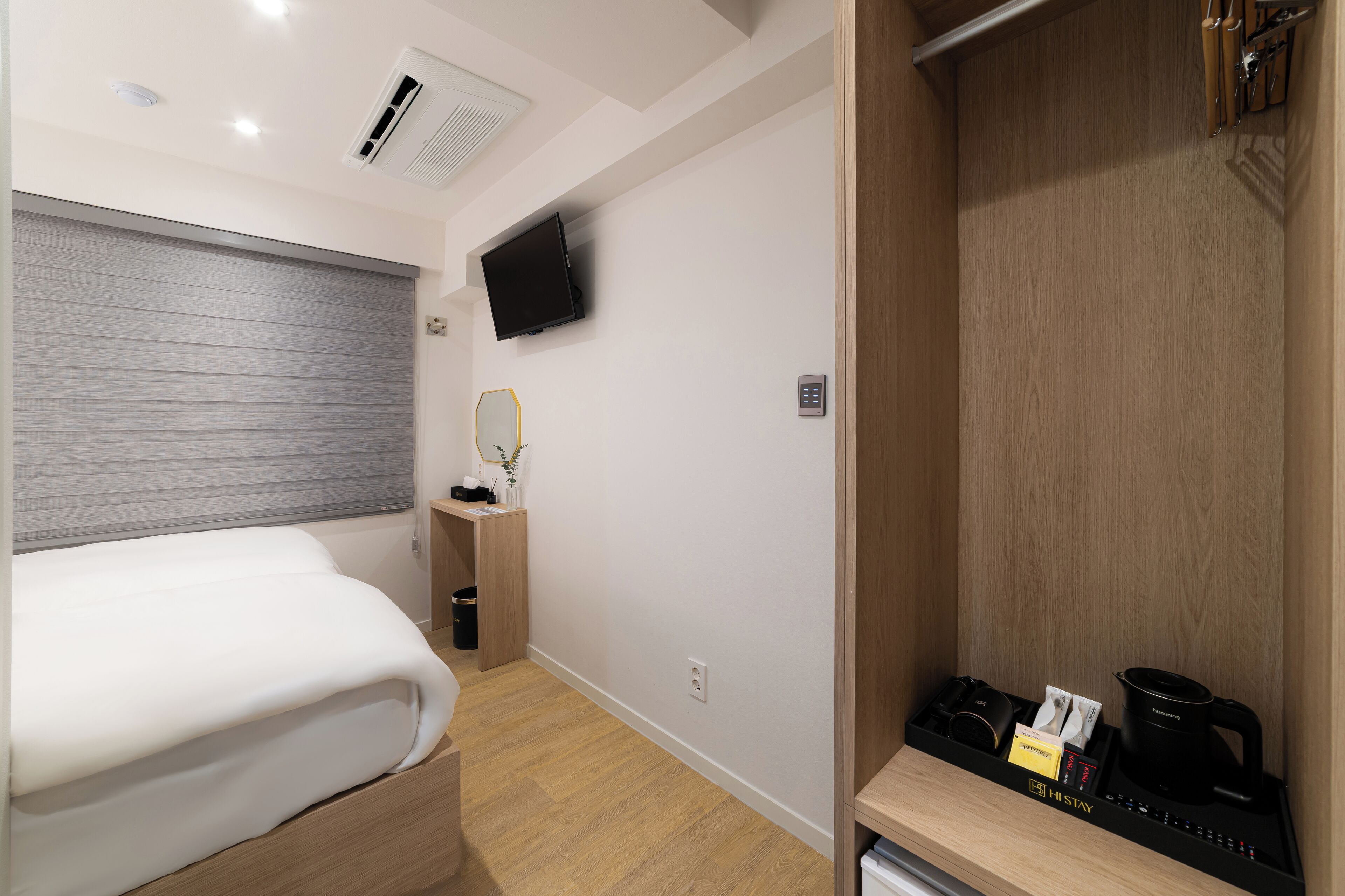 A hotel room with a bed, a TV mounted on the wall, a small bedside table with a mirror, and a wooden closet with a coffee maker and other items.