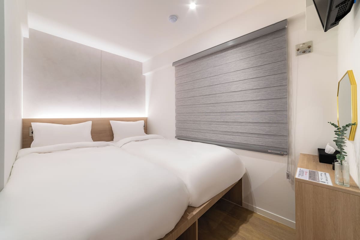 A modern hotel room with a large bed, a bedside table with a mirror, and a window with blinds.