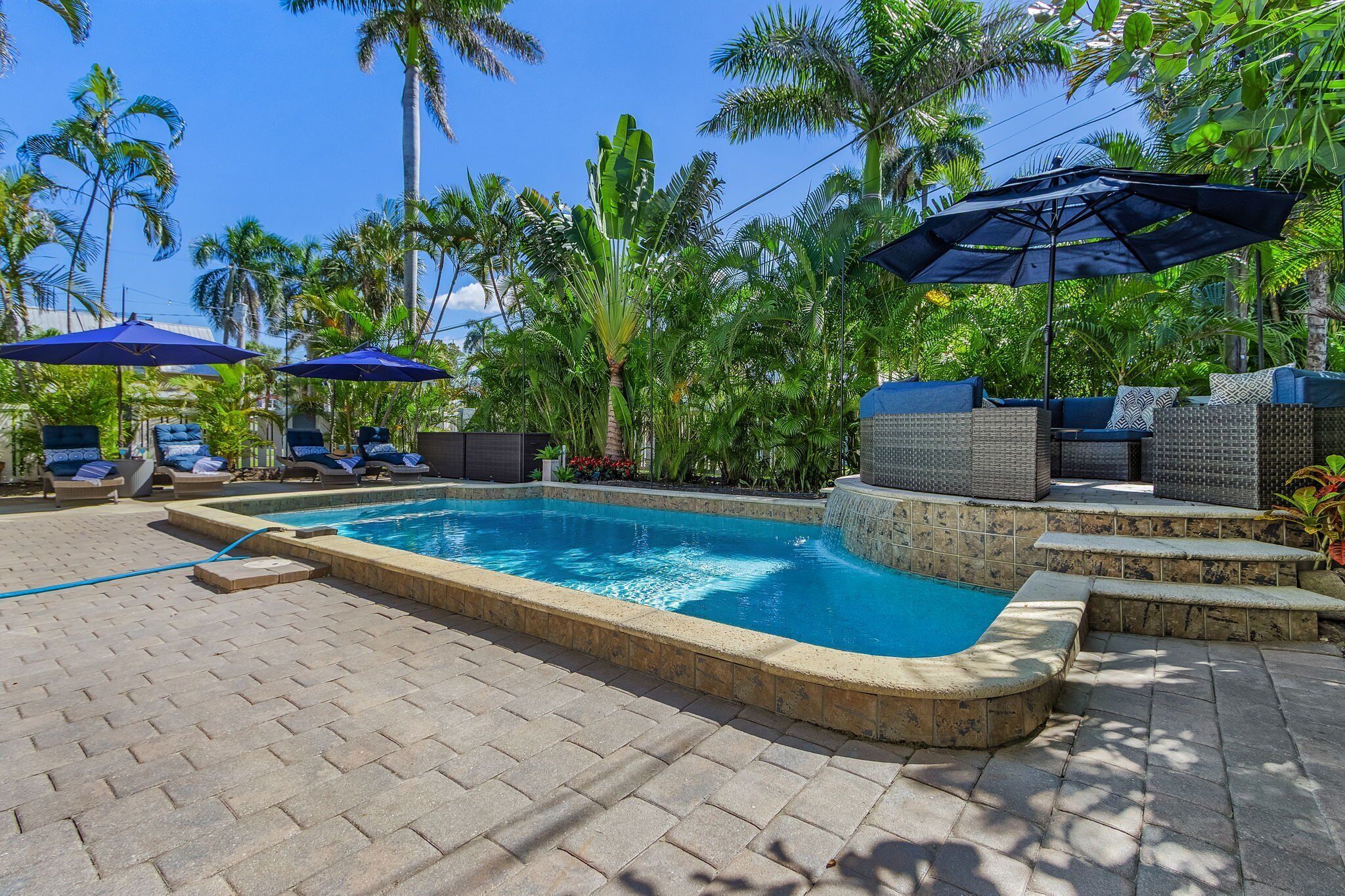 Ein Swimmingpool mit Liegestühlen und Sonnenschirmen, umgeben von Palmen und einer Pflasterterrasse.