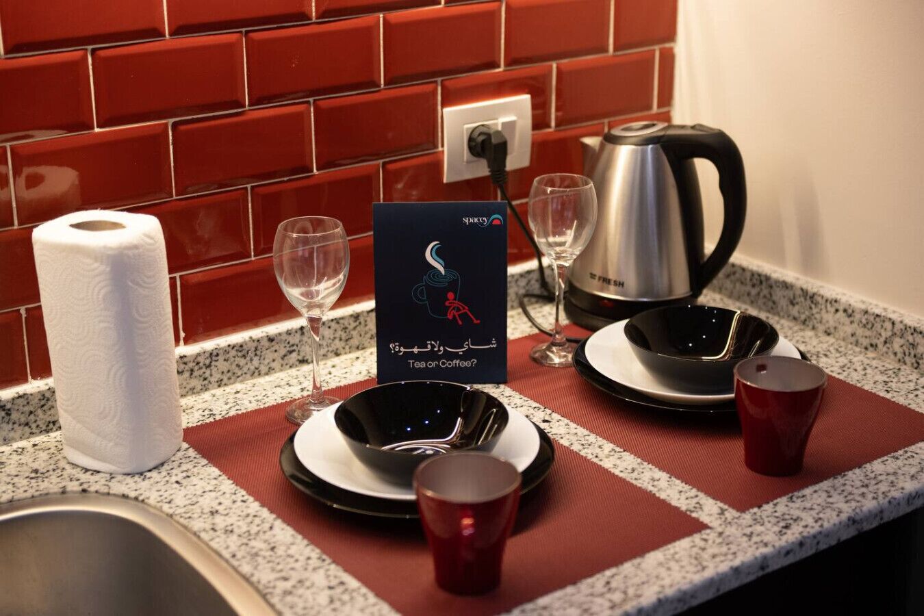 En køkkenbordplade med en kaffemaskine, to glas, et skilt med 'te eller kaffe', en rulle køkkenrulle og en rød kop.