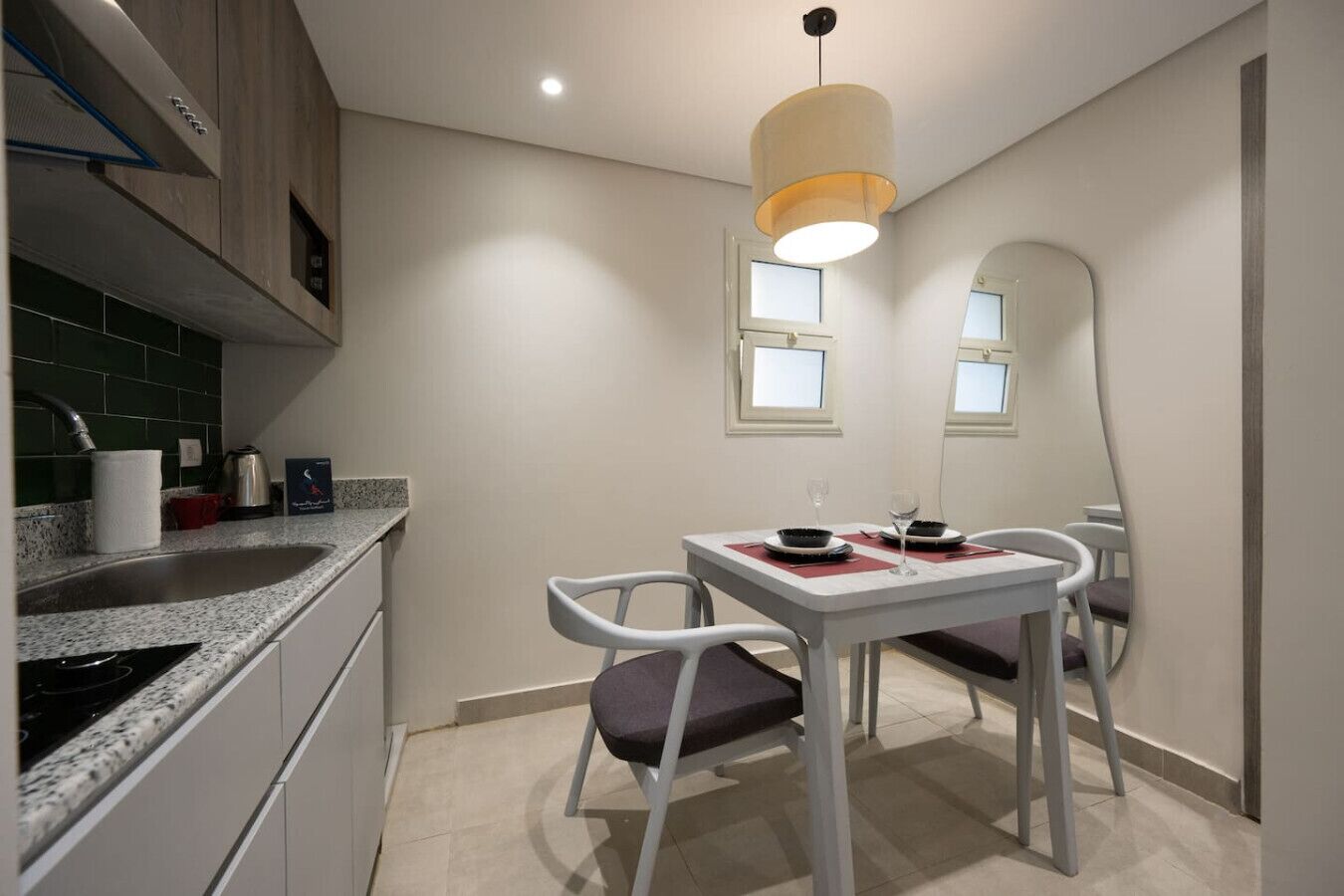 A compact kitchen with a dining area, featuring a granite countertop, a stainless steel sink, a white dining table with two chairs, and a pendant light.