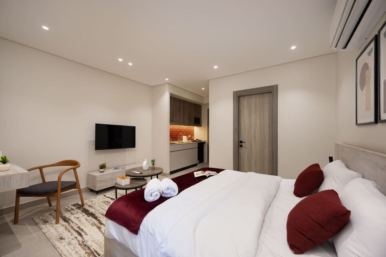 A hotel room with a bed, a desk with a chair, a TV mounted on the wall, and a kitchenette in the background.