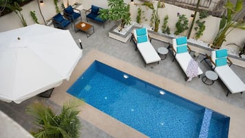Une piscine extérieure, entourée de plantes et dotée d’un parasol blanc, avec des chaises longues et une table à proximité.