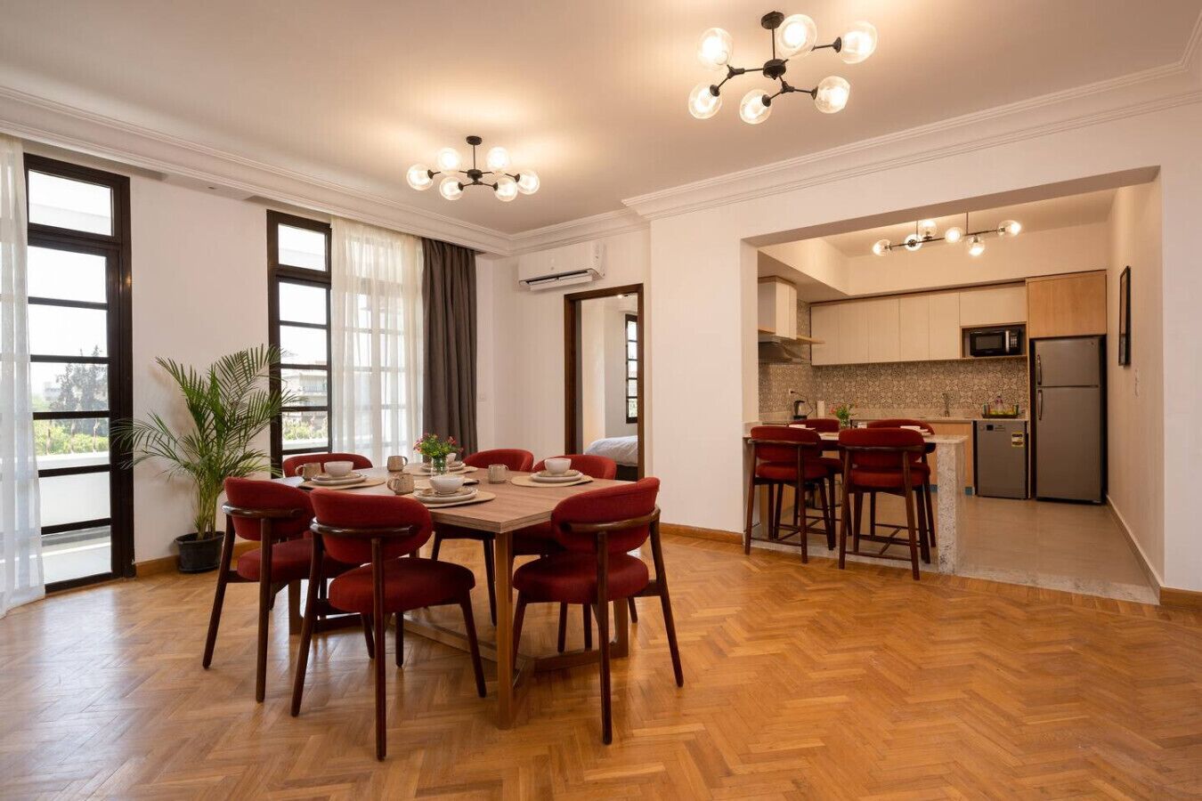A spacious dining area with a wooden herringbone floor, a large wooden table, and red upholstered chairs. There is a potted plant to the left and a kitchen area with modern appliances visible through an open doorway.