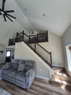Un vaste espace de vie comprenant un canapé gris, un escalier en bois et un ventilateur de plafond.