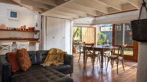 Ein großzügiger Wohnbereich mit einer schwarzen Ledersofa, einem Holzespeisetisch und Stühlen. Ein Küchenbereich mit weißen Schränken und einer hängenden Pfannenträger ist sichtbar.