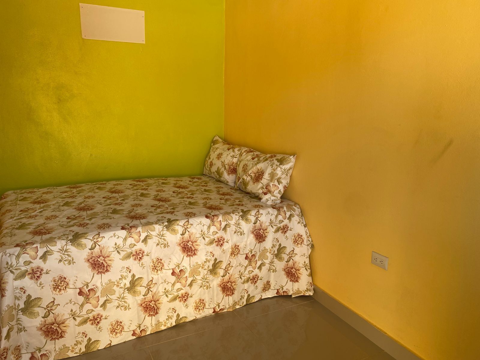 Une petite chambre avec un lit simple, une couvre-lit à motifs floraux, des murs verts et jaunes, et une prise électrique visible sur le mur jaune.