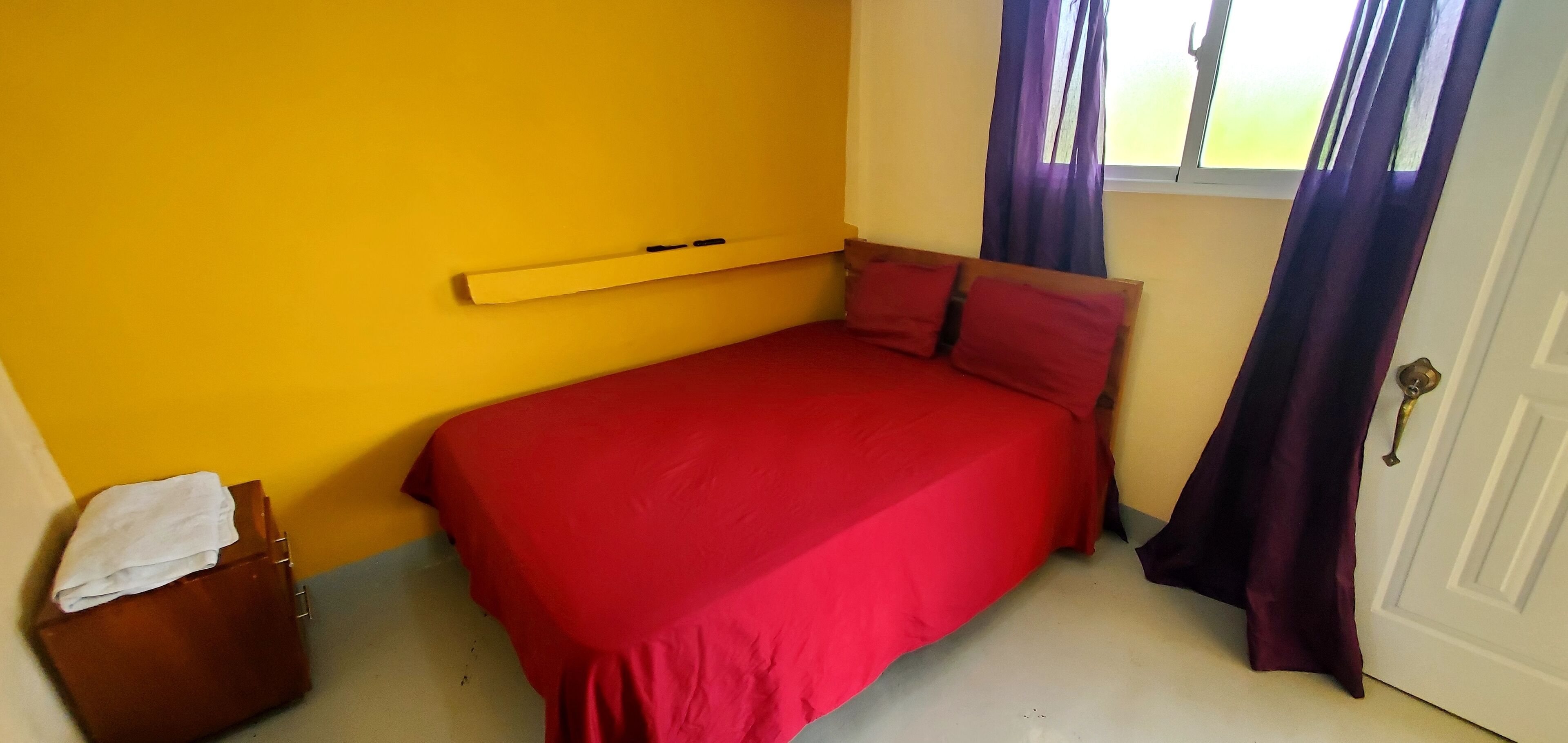 A small, single-bed room with a red bedspread, a wooden headboard, a window with purple curtains, and a door with a brass handle.