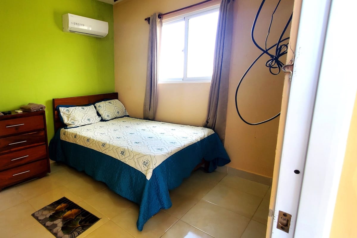A bedroom with a bed, a dresser, a window with curtains, and an air conditioning unit.