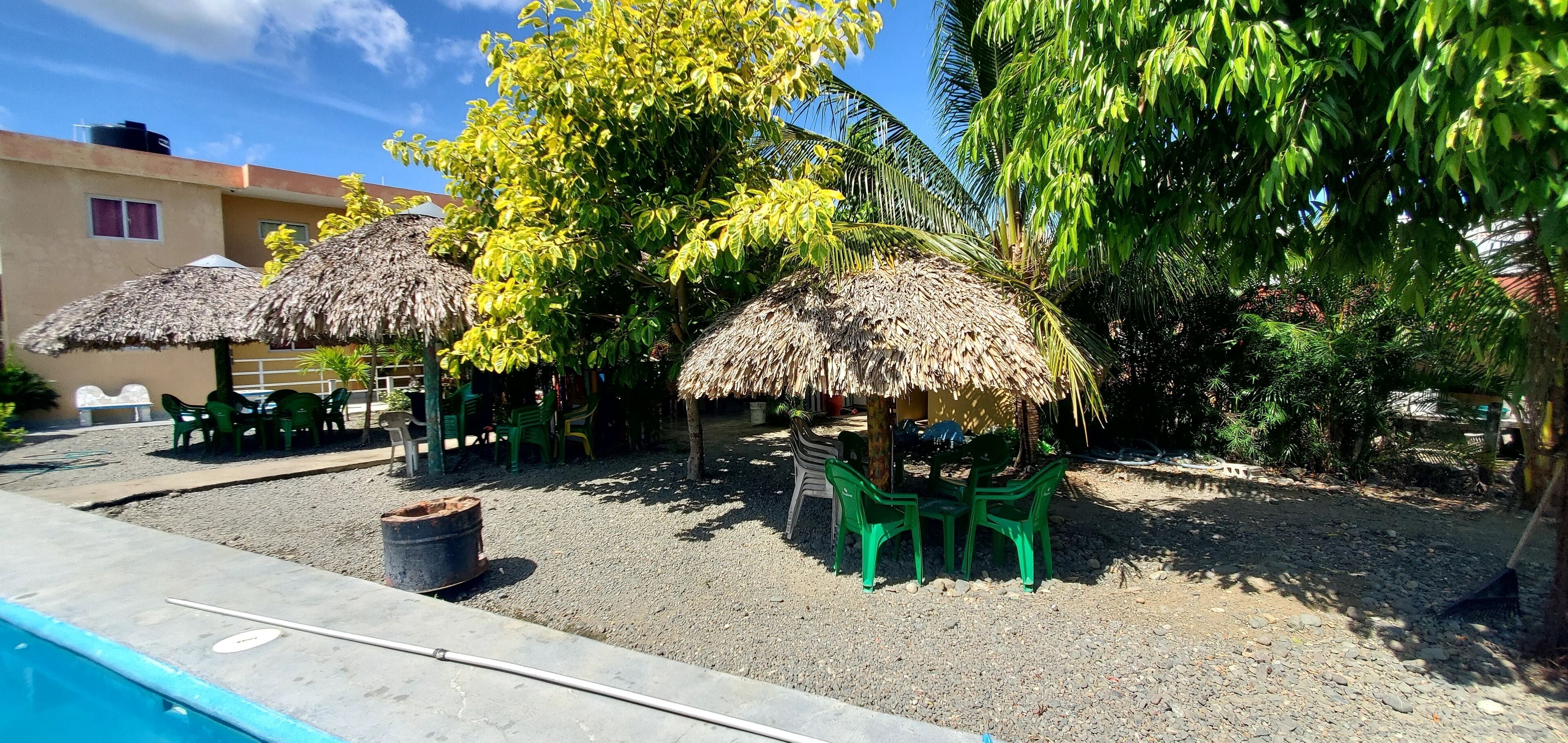 Zona de piscina con sombrillas de paja y sillas de plástico verde.