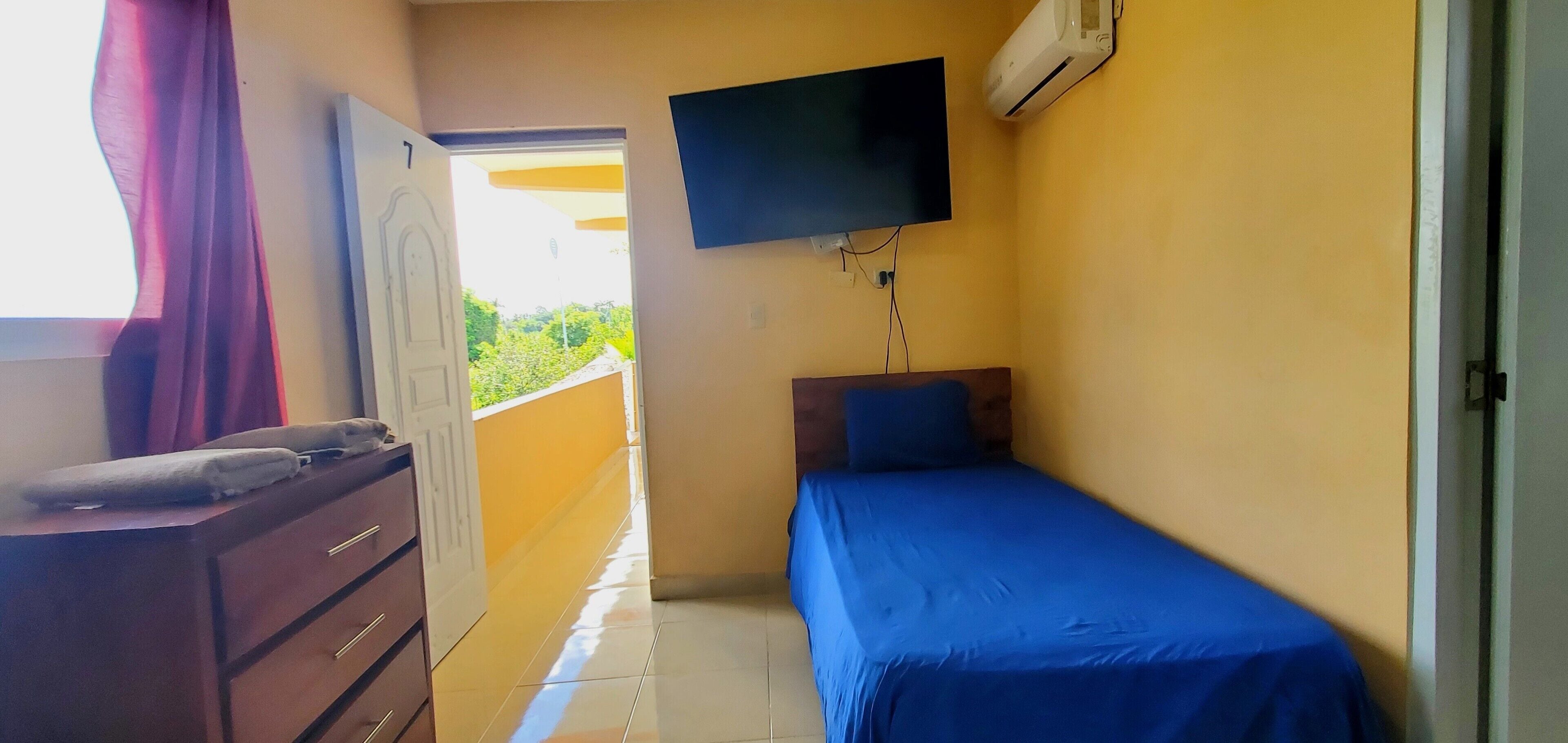 Una habitación pequeña con una cama, un televisor montado en la pared y un ventanal con cortinas.