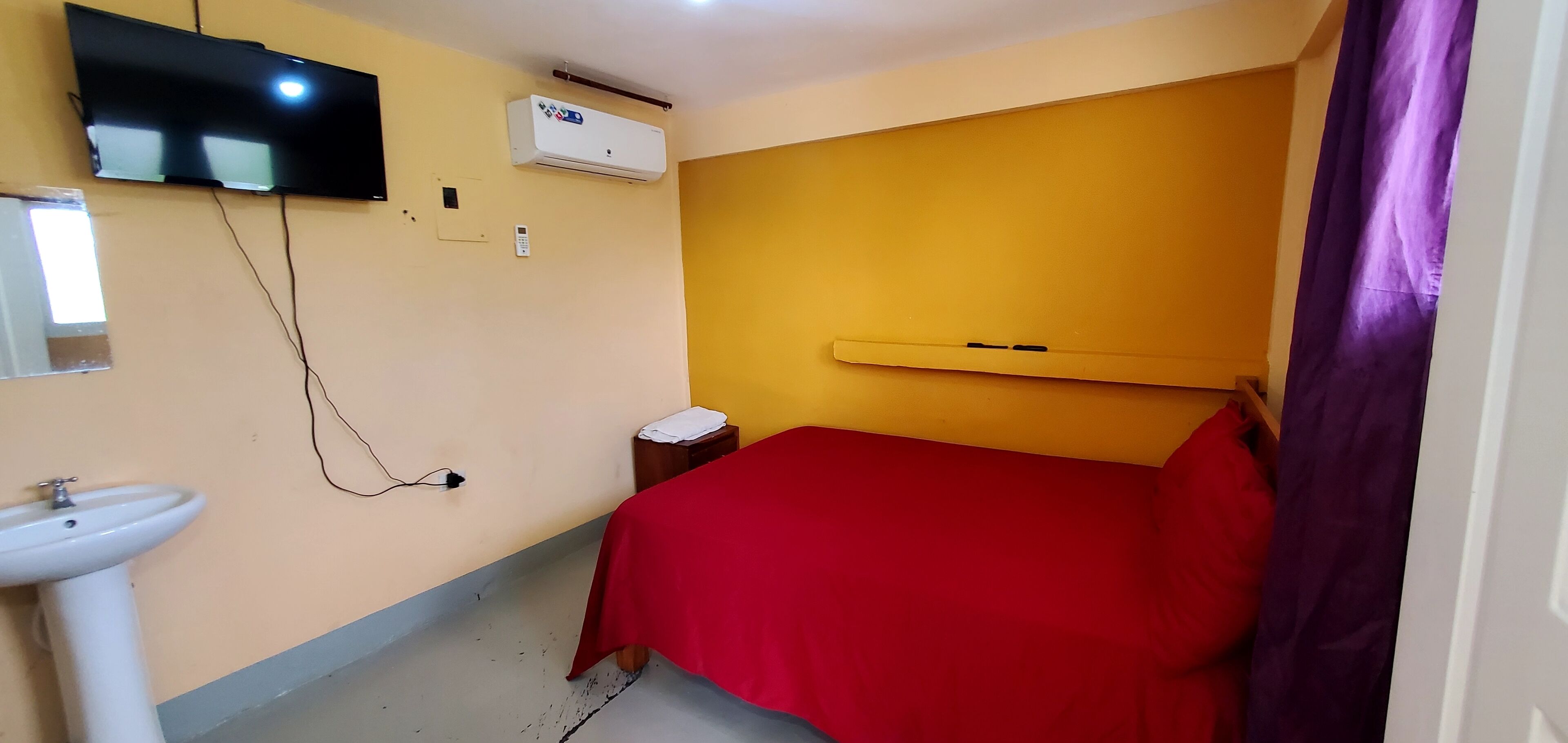 Een kamer met een bed, een aan de muur bevestigde televisie, een airconditioning, een nachtkastje met een lamp en een raam met gordijn.