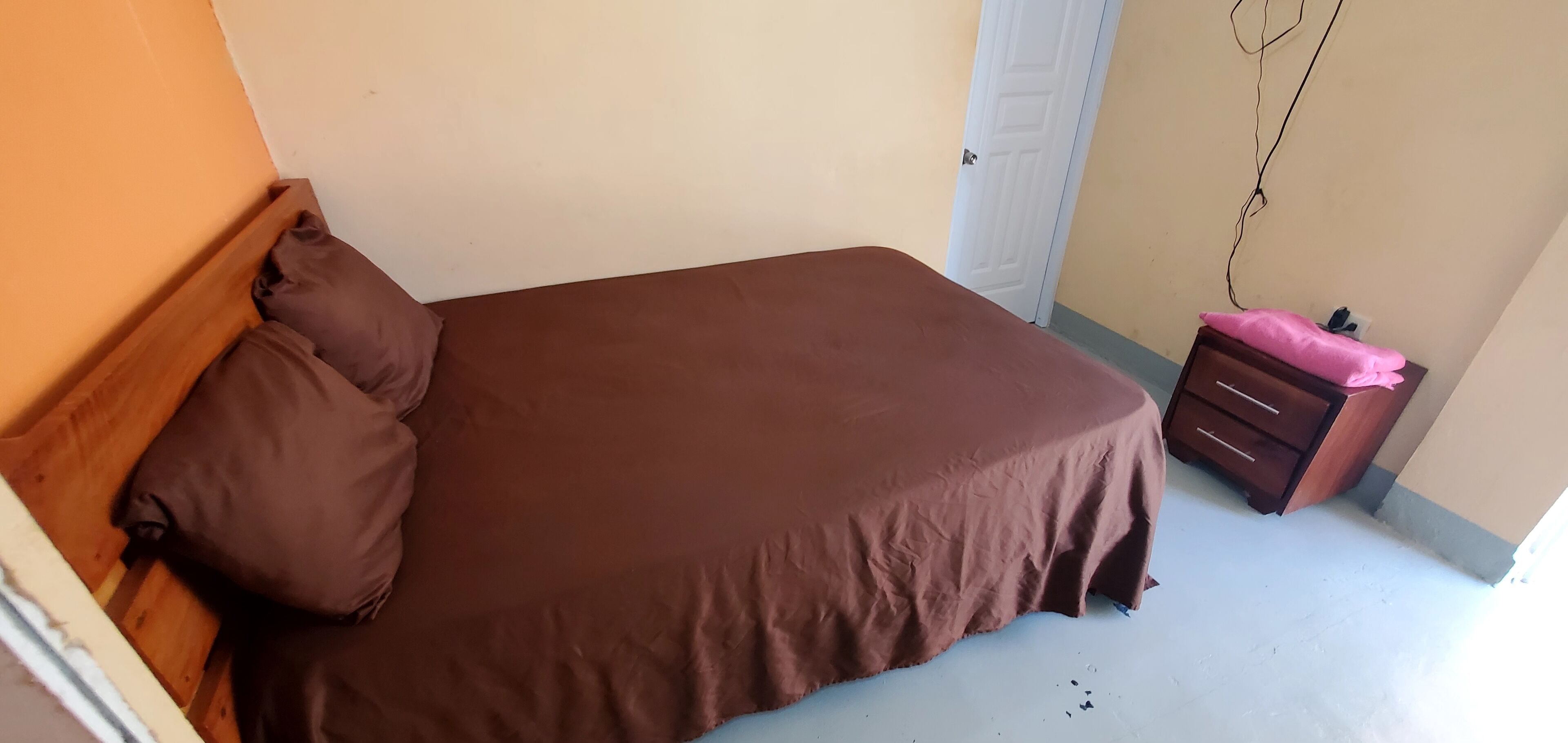 A bedroom with a brown bed, a wooden headboard, a brown pillow, a wooden nightstand with a pink towel, and a white door.