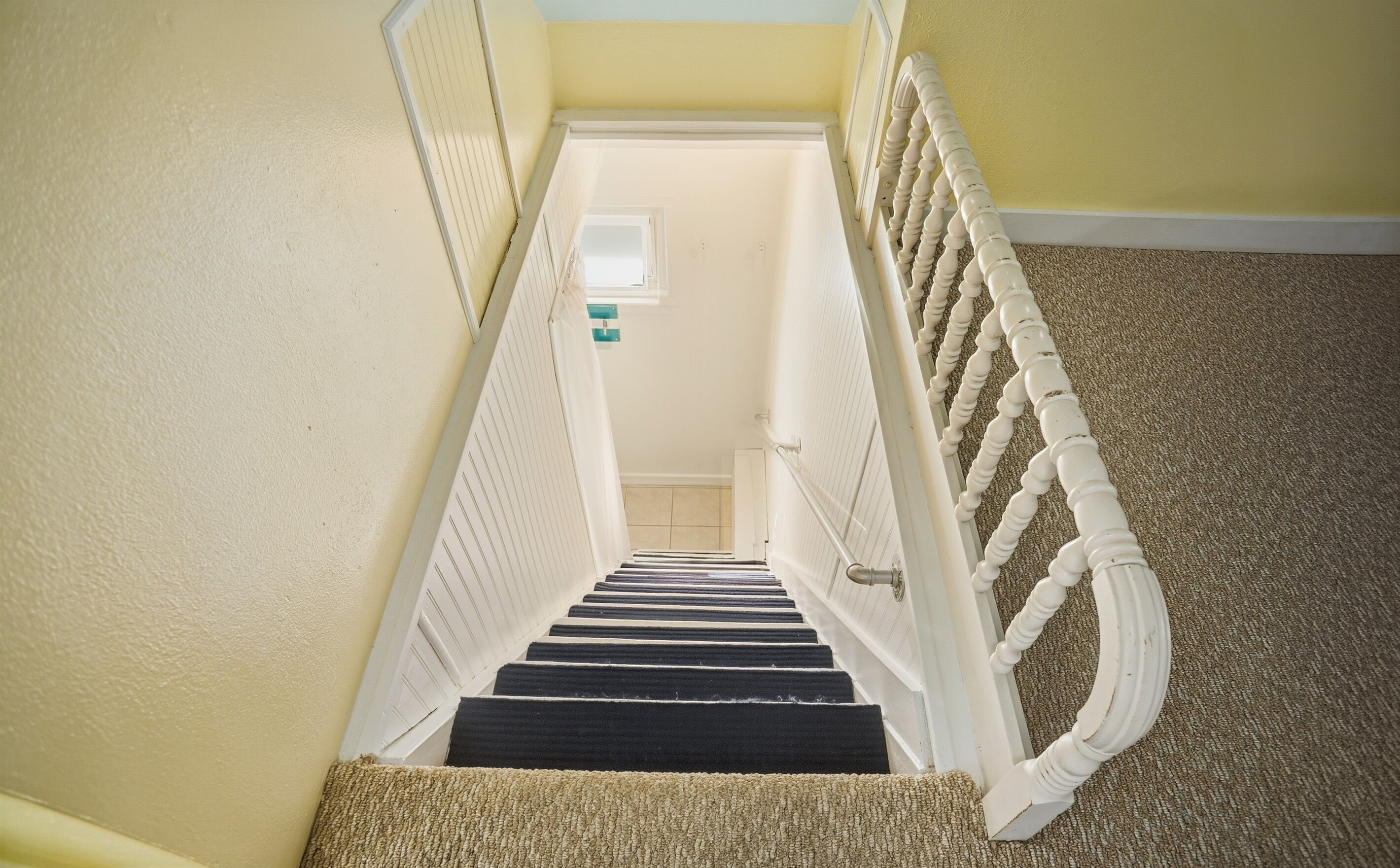 Una escalera alfombrada con balaustres blancos y barandilla, que conduce a una habitación con pared de color claro y un ventanal pequeño.