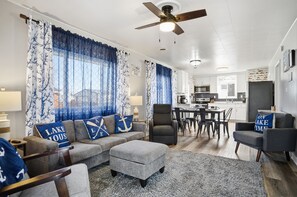 Ein modernes Wohnzimmer mit grauem Sofa, Ottomanen und Sesseln, einem Essbereich mit Tisch und Stühlen sowie einer Küche im Hintergrund.