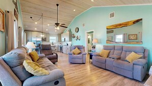 A spacious living room with a large grey sectional sofa, a smaller grey armchair, and a wooden coffee table. The room has light blue walls, wooden flooring, and a ceiling fan. - Pet Friendly, Golf Cart Access, Old Town Location (Port Aransas)