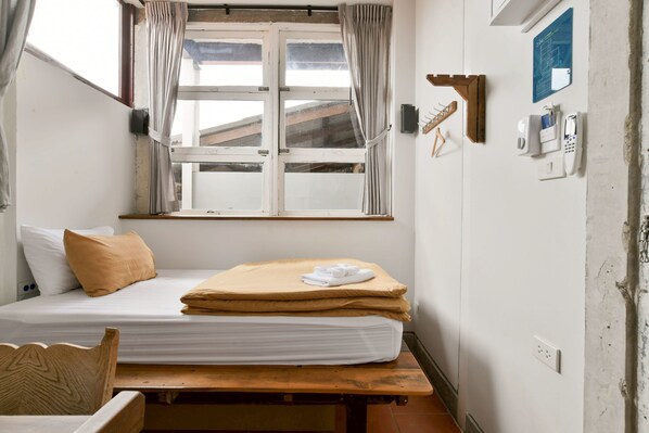 A small, compact room with a single bed, a wooden bench, a window with curtains, a wall-mounted phone, and a bulletin board. - VELAWARIN HOTEL (Warin Chamrap)