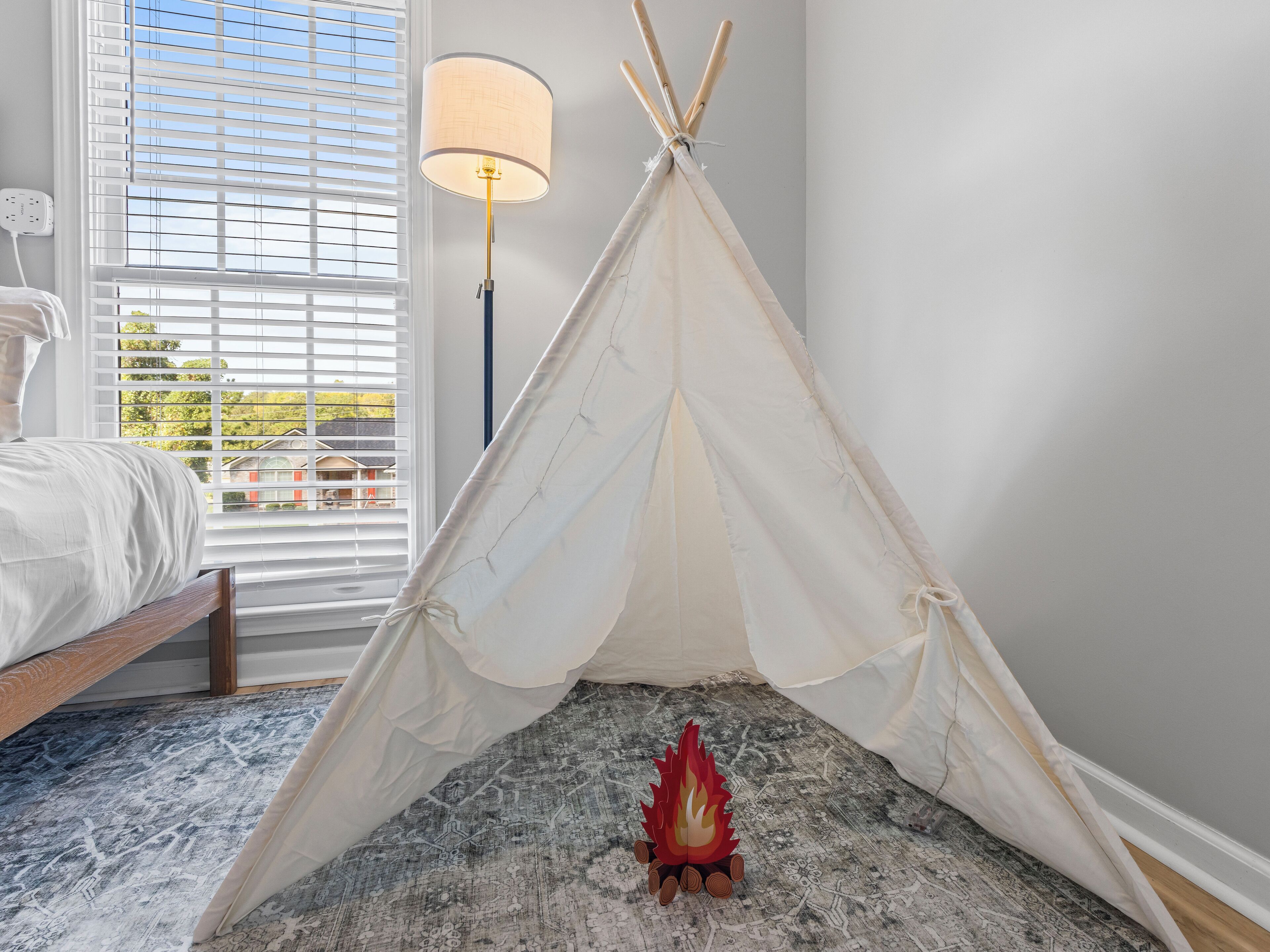 Une chambre avec un lit, une fenêtre avec des stores, une lampe et une tente tipi avec un jouet représentant un feu de camp.